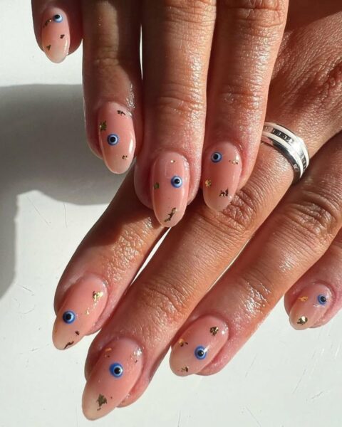 Zdjęcie dłoni ze zbliżeniem na evil eye manicure, czyli długie, owalne paznokcie ozdobione motywem oka proroka. Na palcu serdecznym widać srebrno-białą obrączkę. Dłonie układają się tak, by prezentować manicure. Pada na nie światło słoneczne i lśnią.