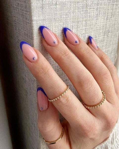 Zdjęcie dłoni z długimi, migdałowymi paznokciami pomalowanymi w stylu french manicure ale z niebieskimi końcówkami oraz ze wzorem oka proroka u nasady. Na palcach widać liczne złote pierścionki.