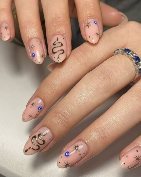 Zdjęcie dłoni ze zbliżeniem na evil eye manicure. Paznokcie są owalne, długie, w kolorze nude i ozdobione wzorami gwiazd, oka proroka oraz złotego węża. Dłonie pozują na białym tle. Na palcu środkowym jednej z dłoni jest srebrny pierścionek z niebieskimi kamieniami.