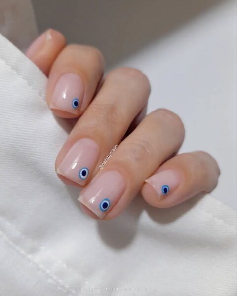 Zdjęcie dłoni z kwadratowymi paznokciami z corner nail art. Paznokcie są pomalowane na kolor nude i ozdobione wzorem oka proroka w rogu paznokcia.