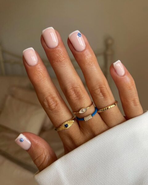 Zdjęcie dłoni z kwadratowymi paznokciami. Są pomalowane w stylu klasycznego french manicure, ale paznokcie kciuka i palca serdecznego zdobi małe oko proroka. Na palcach widać złote pierścionki z niebieskimi motywami.