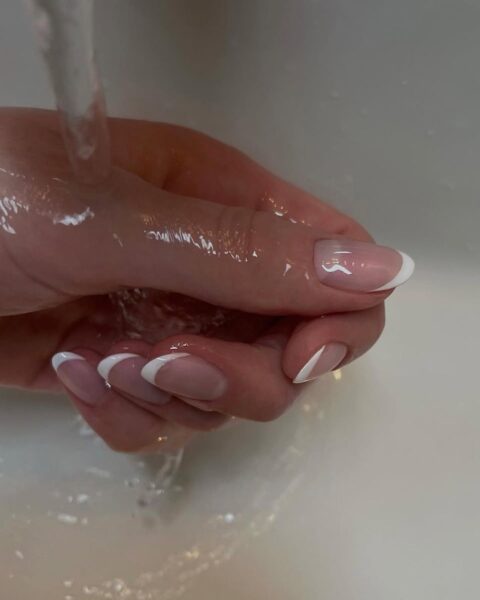 Zdjęcie dłoni ze zbliżeniem na paznokcie, owalne i długie. Zdobi je french manicure dla 50-latek z lśniącym wykończeniem. Dłoń znajduje się pod kranem, leci na nią woda.