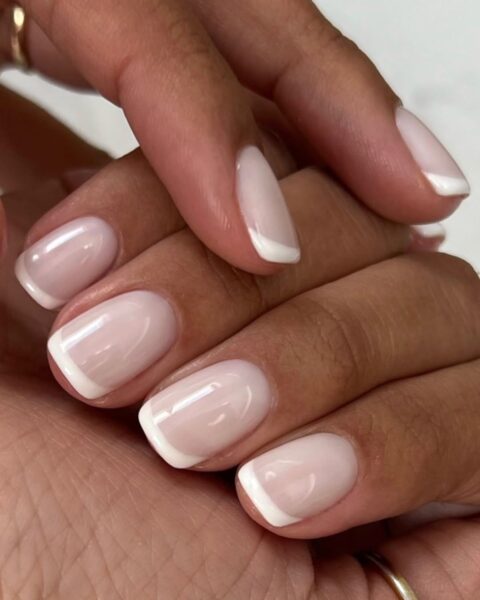 Zdjęcie dłoni ze zbliżeniem na kwadratowe paznokcie z zaokrąglonymi brzegami ze stylizacją french manicure w dwóch odcieniach bieli. Dłonie układają się tak, by prezentować manicure.