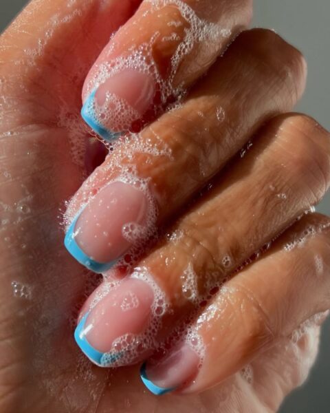 Zdjęcie dłoni ze zbliżeniem na kwadratowe paznokcie z zaokrąglonymi brzegami. Mają niebieskie końcówki w stylu french manicure. Dłonie są pokryte pianą.