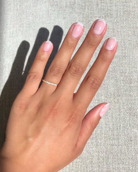 Zdjęcie dłoni ze zbliżeniem na owalne paznokcie w kolorze nude i podwójną, białą końcówką w styli double french manicure. Na palcu serdecznym widać cienki pierścionek z cyrkoniami.