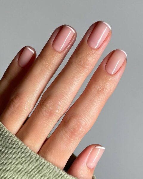 Zdjęcie dłoni ze zbliżeniem na kwadratowe paznokcie z zaokrąglonymi bokami. Są pomalowane w stylu micro french manicure z bardzo cienką kreseczką na brzegu paznokci.