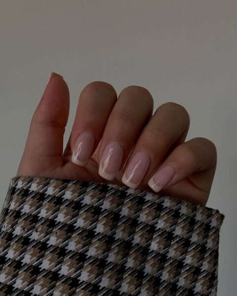 Zdjęcie dłoni z długimi, kwadratowymi paznokciami z french manicure z beżową końcówką na brzegach paznokci. Widać fragment rękawa marynarki we wzór w kratkę.