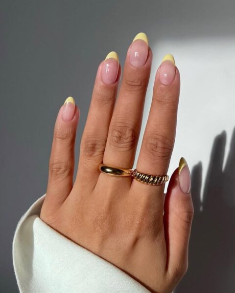 Zdjęcie dłoni ze zbliżeniem na długie, owalne paznokcie pomalowane w stylu french manicure z żółtymi końcówkami. Na palcach widać złote pierścionki.
