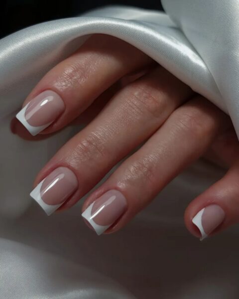 Zdjęcie dłoni ze zbliżeniem na długie, kwadratowe paznokcie. Są pomalowane w stylu french manicure w kolorze białym. Dłoń pozuje na tle białego materiału.