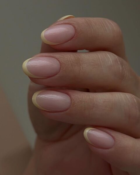 Zdjęcie dłoni ze zbliżeniem na średniej długości, owalne paznokcie z french manicure w kolorze maślanej żółci.