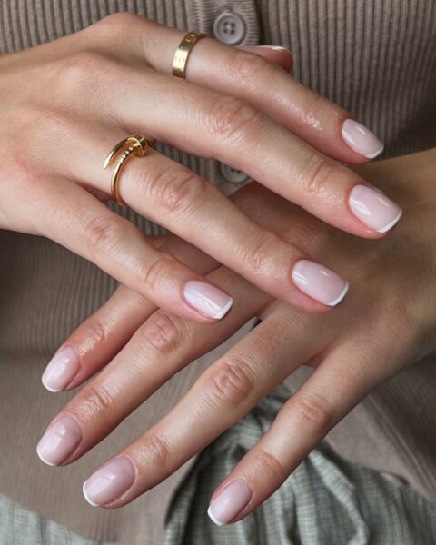 Zdjęcie dłoni ze zbliżeniem na krótkie paznokcie o kształcie zaokrąglonego kwadratu z french manicure w stylu old money. Na palcach znajdują się złote pierścionki marki Cartier. Dłonie pozują na tle beżowego kardiganu.