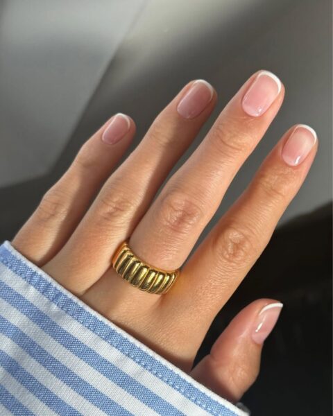Zdjęcie dłoni z krótkimi, kwadratowymi paznokciami z micro french manicure w stylu old money. Na środkowym palcu znajduje się złoty pierścionek. Widać fragment koszuli w biało-niebieskie paski.