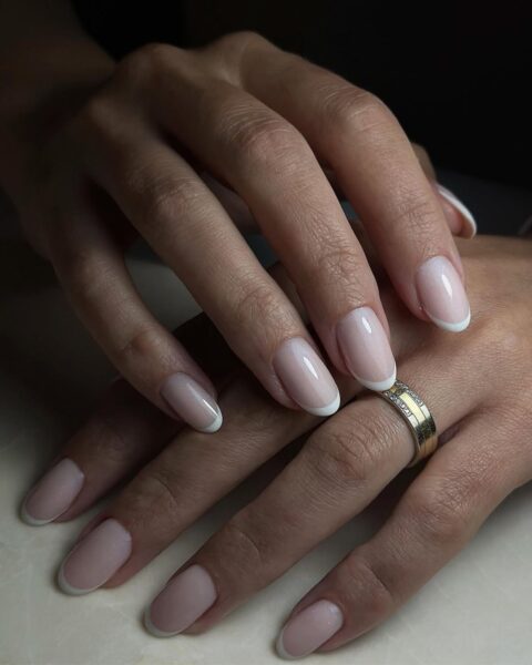 Zdjęcie dłoni z owalnymi, średniej długości paznokciami pomalowanymi w stylu french manicure. Na palcu serdecznym widać złotą obrączkę. Dłonie układają się tak, by prezentować paznokcie.