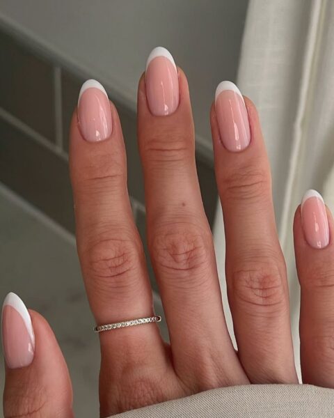 Zdjęcie dłoni ze zbliżeniem na średniej długości, owalne paznokcie pomalowane w stylu french manicure. Na palcu wskazującym znajduje się cienka, złota obrączka z cyrkoniami.