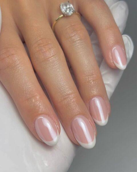 Zdjęcie dłoni ze zbliżeniem na średniej długości, owalne paznokcie pomalowane w stylu french manicure z chromowanym wykończeniem. Na palcu serdecznym znajduje się złoty pierścionek z dużym brylantem. Dłoń opiera się o dłoń w białej rękawiczce stylistki paznokci.