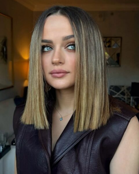 Zdjęcie kobiety w blond, prostych włosach do ramion w stylu sleek bob. Ubrana jest w brązową, skórzaną kamizelkę. Pozuje w salonie piękności. Patrzy w bok.
