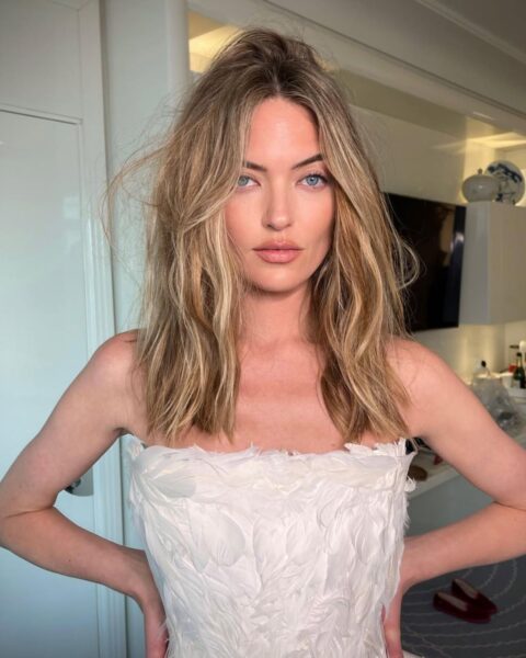 Zdjęcie modelki w jasnych, blond włosach do ramion, lekko rozwianych, naturalnych, falowanych. Ubrana jest sukienkę gorsetową ozdobioną białymi piórami.