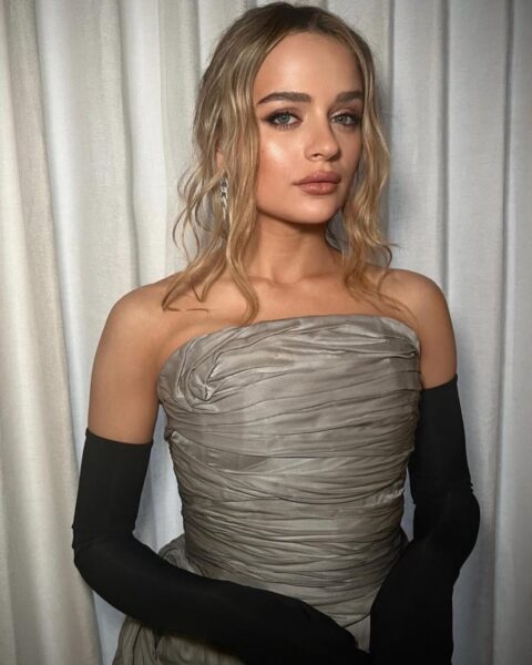 Zdjęcie aktorki Joey King w stylizacji na wesele. Ma fryzurę dla dojrzałej kobiety, czyli lekko falowane blond włosy spięte w luźny kok oraz wypuszczone przy twarzy pasemka. Ubrana jest w marszczoną, szarą sukienkę odkrywającą ramiona oraz czarne, długie rękawiczki. Pozuje na tle kremowej zasłony.