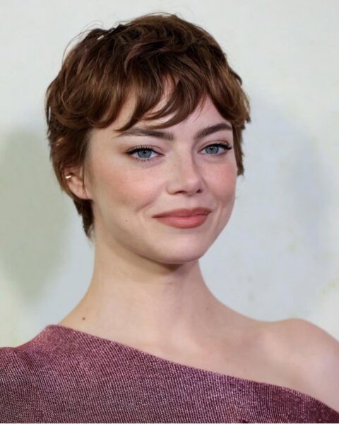 Zdjęcie aktorki Emmy Stone w krótkim pixie cut w kolorze kasztanowym. Aktorka pozuje na ściance. Ubrana jest w różową sukienkę o asymetrycznym dekolcie.