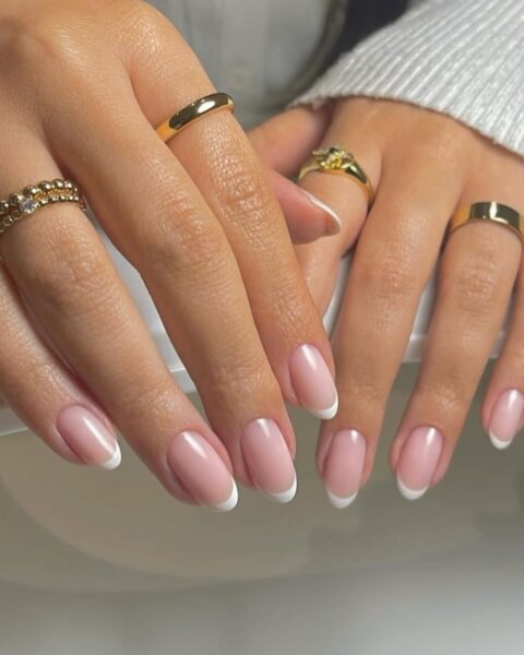 Zdjęcie dłoni z długimi, owalnymi paznokciami z różowym french manicure. Na palcach znajduje się kilka złotych pierścionków. Dłonie pozują tak, by prezentować manicure skierowane w dół.