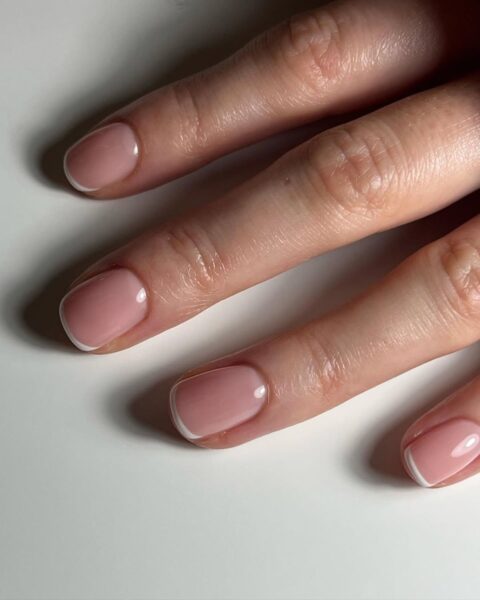 Zdjęcie dłoni ze zbliżeniem na krótkie, owalne paznokcie z french manicure. Dłoń pozuje oparta o biały blat.