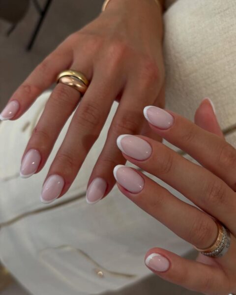 Zdjęcie dłoni ze zbliżeniem na owalne, długie paznokcie ze szklanym francuskim manicure. Na palcach widać złote pierścionki. Dłonie pozują na tle białej sukienki i kremowej bluzki.