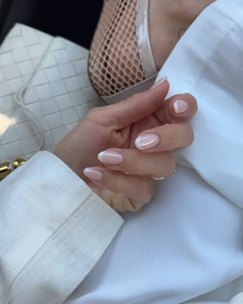 Zdjęcie dłoni ze stylizacją glass french manicure. Widać kobietę, która zrobiła sobie zdjęcie z góry, siedząc w samochodzie. Prezentuje manicure, ubrana jest w białe spodnie i siateczkowe baleriny. Opiera nogę o kolano. Widać fragment beżową torebkę plecioną.