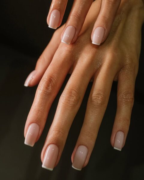 Zdjęcie dłoni ze zbliżeniem na kwadratowe paznokcie z lśniącym french manicure. Dłonie pozują na czarnym tle tak, by prezentować manicure.