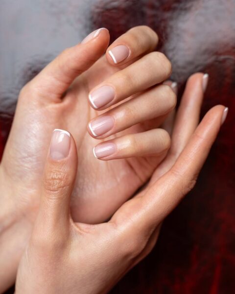 Zdjęcie dłoni ze zbliżeniem na krótkie, kwadratowe paznokcie z french manicure. Dłonie pozują tak, by prezentować manicure i układają się na tle bordowym, lśniącym tle.
