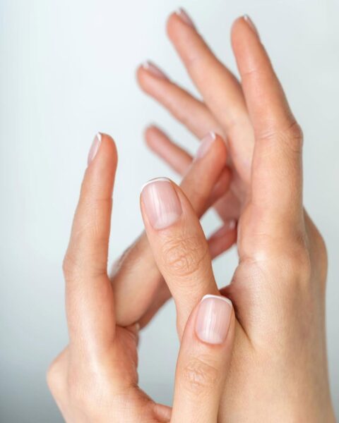 Zdjęcie dłoni ze zbliżeniem na krótkie, kwadratowe paznokcie z zaokrąglonymi bokami i z francuskim manicure. Dłonie układają się tak, by prezentować manicure.