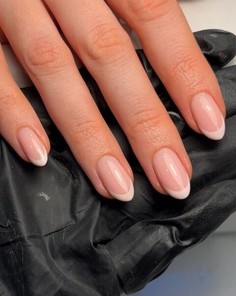 Zdjęcie dłoni z długimi, owalnymi paznokciami z beżowym french manicure. Dłoń opiera się o dłoń stylistki paznokci w czarnej rękawiczce.