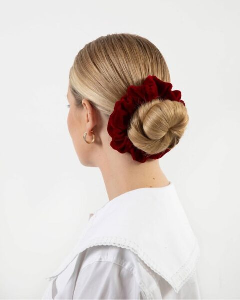 Zdjęcie kobiety stojącej tyłem. Ma blond włosy zwinięte w ślimaka i upięte welurową frotką w kolorze bordowym. Ubrana jest w białą koszulę z szerokim kołnierzem. W uszach ma złote kolczyki.