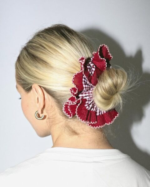 Zdjęcie kobiety stojącej tyłem. Ma blond włosy związane w kok i spięte dużą, bordowo-białą scrunchie w kratkę i z falbanką marki Staise. Ubrana jest w biały top i ma w uszach złote kolczyki kółka.