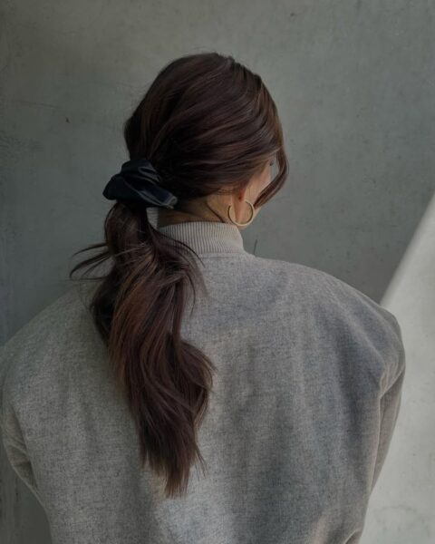 Zdjęcie kobiety stojącej tyłem. Ubrana jest w szarą kurtkę. Ma ciemne włosy związane w niski kok za pomocą czarnej scrunchie. Fryzury tego typu są proste i wygodne. W uszach ma złote, okrągłe kolczyki. Kobieta pozuje na tle szarej ściany.