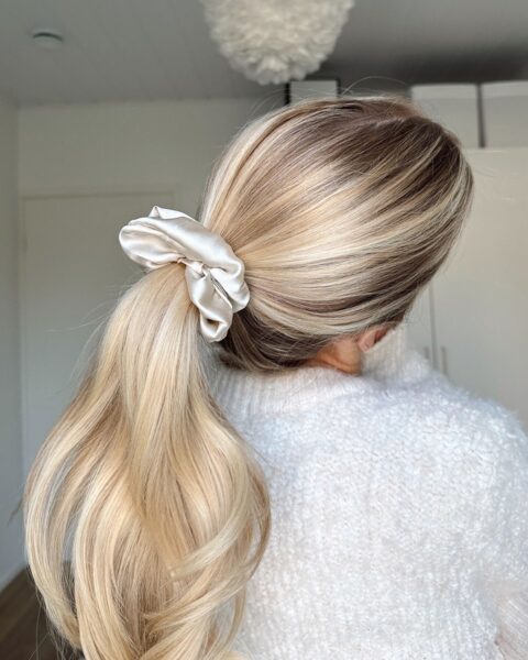 Zdjęcie kobiety stojącej tyłem. Ma blond włosy związane w wysoki koński ogon z kremową frotką, jako przykład scrunchie fryzury. Ubrana jest w biały sweter. Pozuje w sypialni na przeciwko szafy białej, widać też żyrandol.