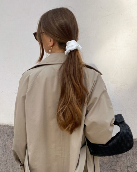 Zdjęcie kobiety stojącej tyłem. Ma brązowe włosy związane w niski kok z pomocą białej scrunchie. Ubrana jest w beżowy płaszcz typu trencz, a na ramieniu ma czarną torebkę Miu Miu.