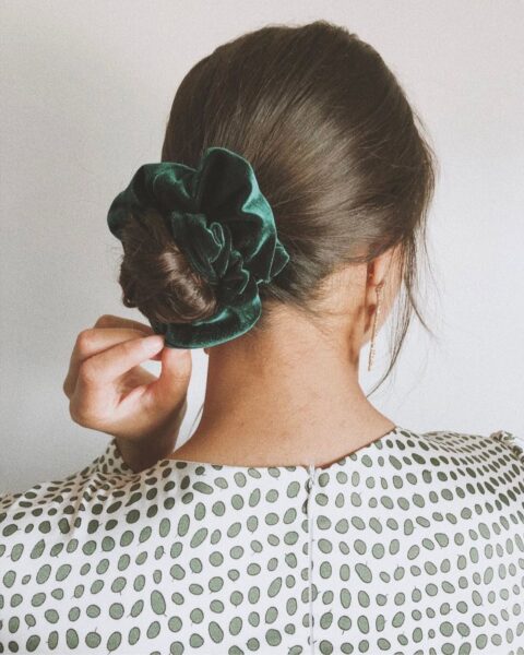 Zdjęcie kobiety stojącej tyłem. Ma brązowe włosy związane w kok za pomocą zielonej scrunchie. Ubrana jest w białą bluzkę w zielone groszki.