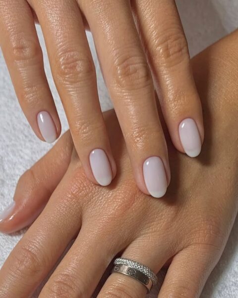 Zdjęcie dłoni ze zbliżeniem na długie, owalne paznokcie. Są pomalowane w stylu french manicure w kolorze mlecznej bieli. Na palcu serdecznym jednej z dłoni znajdują się dwie obrączki. Dłonie pozują na tle białego ręcznika.