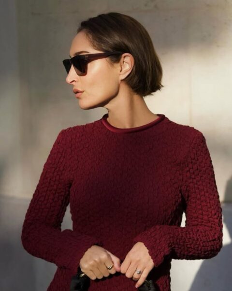 Zdjęcie kobiety, Francuzki ubranej w bordową, dopasowaną sukienkę z długimi rękawami. Ma ciemne włosy do żuchwy, w stylu jawline bob. Na nosie ma okulary przeciwsłoneczne, a w rękach czarną torebkę. Ma też wyrazisty makijaż ust.