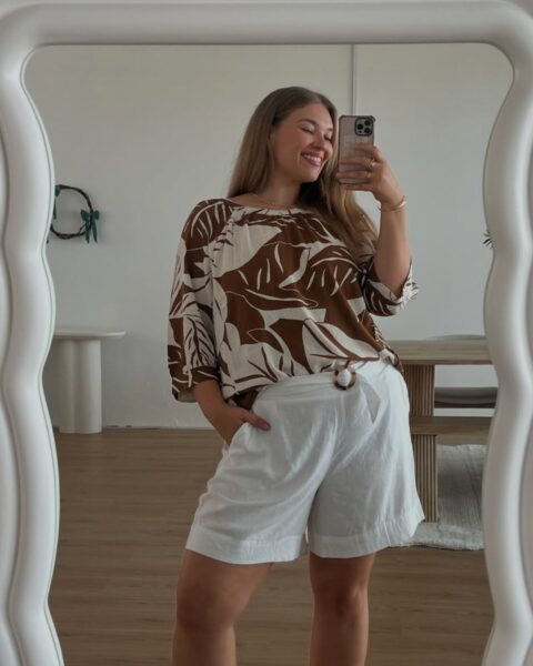Zdjecie kobiety plus size, która znalazła sposób na to, jak ukryć brzuch w letnich stylizacjach: ubrała białe szerokie szorty oraz luźną bluzkę w kolorze kremowo-brązowym ze wzorem kwiatów. Robi sobi selfie w lustrze. Ma długie, blond włosy rozpuszczone na ramiona. Za nią widac pomieszczenie z panelami na podłodze i z białymi ścianami.