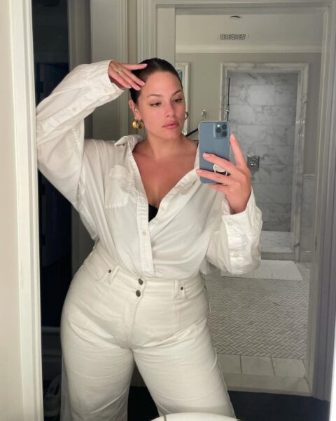 Zdjecie kobiety plus size, która pozuje w łazience i robi sobie selfie w lustrze. Ubrana jest w białe spodnie z wysokim stanem, które maskują jej duży brzuch. W pasie ma włożoną białą koszulę. Włosy ciemne, związane gładko z tyłu. Ma w uszach złote kolczyki.