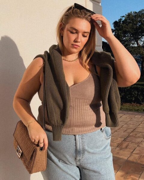 Zdjecie kobiety plus size w letniej stylizacji. Ma na sobie błękitne spodnie jeansowe, beżowy, dzianinowy top na ramiączkach, a na to ma brązowy sweter. Na ramieniu ma brązową, plecioną torebkę na łańcuszku. Na głowie ma okulary przeciwsłoneczne. Ma długie, blond włosy.