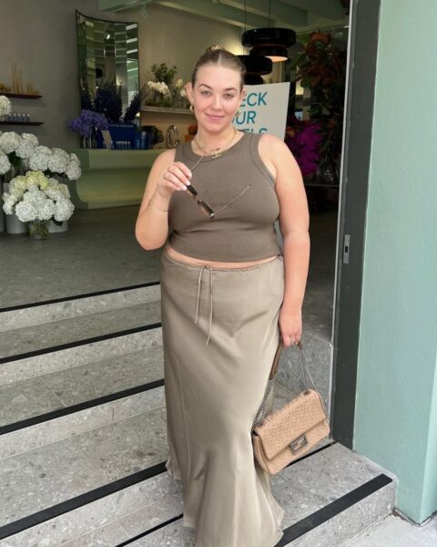 Zdjecie kobiety plus size. Jest ubrana w spódnicę maxi, satynową, w kolorze khaki. Do tego dobrała top w kolorze khaki bez ramiączek. W ręce ma brązową torebkę. Ma jasne, blond włosy związane gładko z tyłu. Pozuje przy wejściu do kwiaciarni, na schodach. W ręce trzyma okulary przeciwsłoneczne.