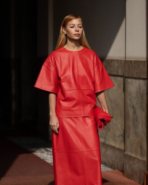 Zdjęcie kobiety ubranej w czerwony komplet: T-shirt oversize oraz ołówkową sukienkę maxi. Do tego ma czerwoną torebkę oraz białe japonki na obcasie. Ma długie, rude włosy, proste, rozpuszczone na ramiona. Pozuje przy bramie, przy wejściu do zabytkowego budynku.