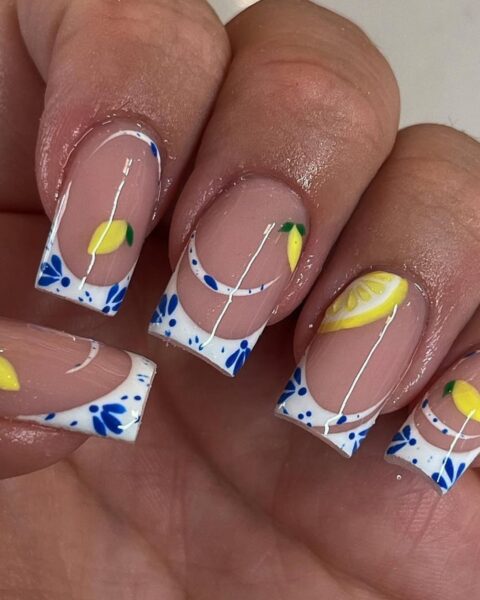 Zdjęcie dłoni ze zbliżeniem na długie, kwadratowe paznokcie ze stylizacją french manicure, ale zamiast białego paska jest pasek z niebieskimi zdobieniami w stylu greckim, a do tego na płytkach paznokci pojawiają się wzory cytryn.
