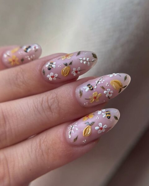 Zdjęcie dłoni ze zbliżeniem na długie, owalne paznokcie pomalowane na kolor pink nude i ozdobione wzorem kwiatów oraz cytryn w artystycznym stylu nail art.