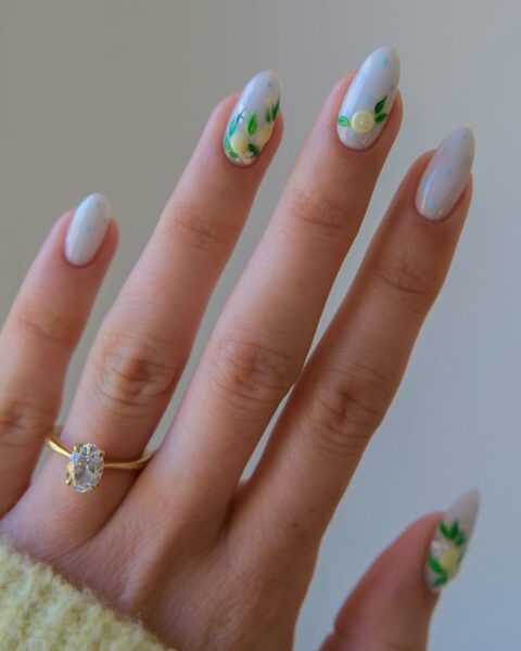 Zdjęcie dłoni ze zbliżeniem na długie paznokcie w kształcie migdała w stylu lemon nails. Są pomalowane na kolor błękitny oraz ozdobione wzorem cytryn. Na palcu serdecznym widać złoty pierścionek z brylantem.