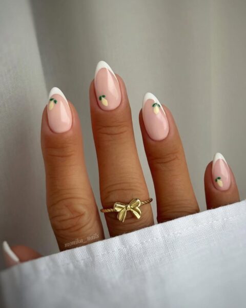 Zdjęcie dłoni ze zbliżeniem na migdałowe, długie paznokcie z french manicure i wzorem cytryn. Na palcu środkowym widać złoty pierścionek z motywem kokardki.