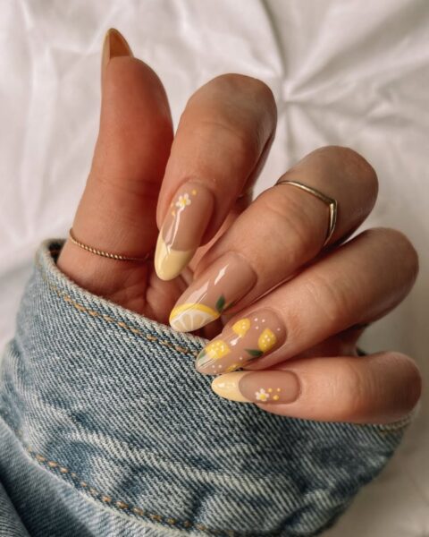 Zdjęcie dłoni ze zbliżeniem na długie, migdałowe paznokcie z żółtym french manicure i wzorem cytryn. Na palcach znajduje się kilka pierścionków. Widać fragment jeansowego rękawa. Dłoń pozuje na tle białego materiału.