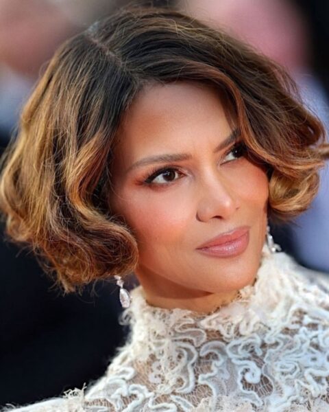 Zdjęcie aktorki Halle Berry w koronkowej, białej sukience ze stójką. Ma opalizujący, odmładzający makijaż dla 40-latki z ocieplającym bronzerem na twarzy. Ma krótkie, ciemne włosy ścięte na boba. W uszach wiszą jej kolczyki z kryształami.